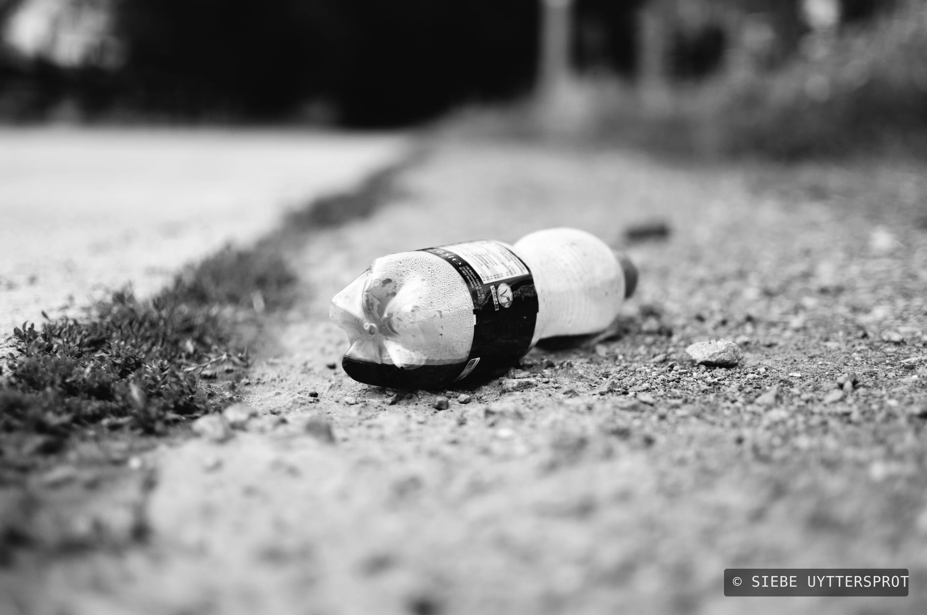 A crushed plastic drink bottle lying on gravel at the edge of a road, condensation beaded on the inside of the plastic.